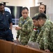 A group of military personnel from multiple nations are gathered around laptops on a wooden table in what appears to be a briefing or operations room. One soldier in camouflage uniform is pointing at a laptop screen while others lean in to observe, suggesting a collaborative training or planning exercise. A large display screen showing a logo or emblem is visible in the background.