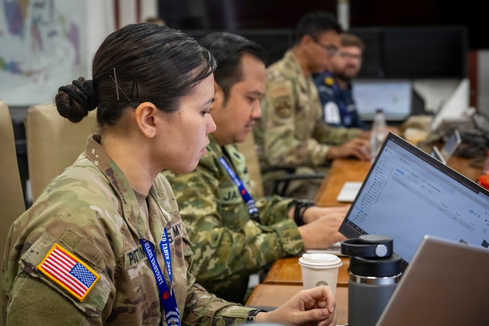 Military personnel in camouflage uniforms are seated at a conference table working on laptops in what appears to be a joint operations or planning environment. The foreground shows a U.S. Army soldier wearing an American flag patch and a blue lanyard, alongside other uniformed individuals. The setting suggests a multinational military exercise or coordination meeting.
