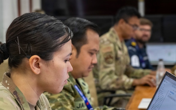 Military personnel in camouflage uniforms are seated at a conference table working on laptops in what appears to be a joint operations or planning environment. The foreground shows a U.S. Army soldier wearing an American flag patch and a blue lanyard, alongside other uniformed individuals. The setting suggests a multinational military exercise or coordination meeting.
