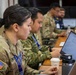 Military personnel in camouflage uniforms are seated at a conference table working on laptops in what appears to be a joint operations or planning environment. The foreground shows a U.S. Army soldier wearing an American flag patch and a blue lanyard, alongside other uniformed individuals. The setting suggests a multinational military exercise or coordination meeting.