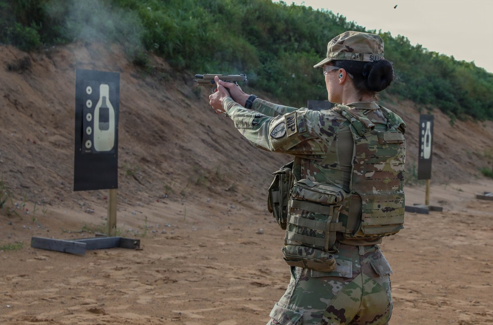 A female soldier in U.S. Army OCP (Operational Camouflage Pattern) uniform is actively firing a pistol at a military shooting range. She is wearing full combat gear including a plate carrier vest, eye protection, and ear protection. A spent casing is visible in the air and smoke is present near the muzzle, indicating the weapon has just been discharged.