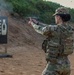 A female soldier in U.S. Army OCP (Operational Camouflage Pattern) uniform is actively firing a pistol at a military shooting range. She is wearing full combat gear including a plate carrier vest, eye protection, and ear protection. A spent casing is visible in the air and smoke is present near the muzzle, indicating the weapon has just been discharged.