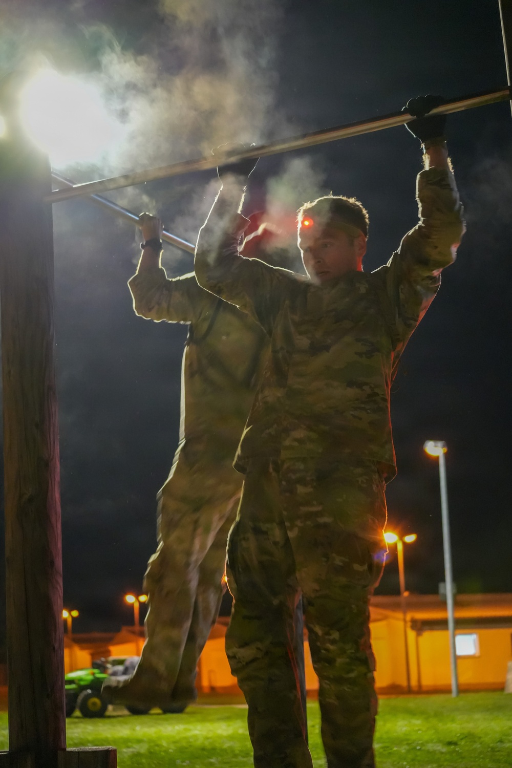 Two military personnel in camouflage uniforms are performing pull-ups or hanging from a horizontal bar at night, illuminated by artificial lighting. The scene appears to take place on a military installation or training facility, with buildings and light poles visible in the background. Visible breath vapor or mist surrounds the subjects, suggesting cold ambient temperatures.