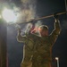 Two military personnel in camouflage uniforms are performing pull-ups or hanging from a horizontal bar at night, illuminated by artificial lighting. The scene appears to take place on a military installation or training facility, with buildings and light poles visible in the background. Visible breath vapor or mist surrounds the subjects, suggesting cold ambient temperatures.