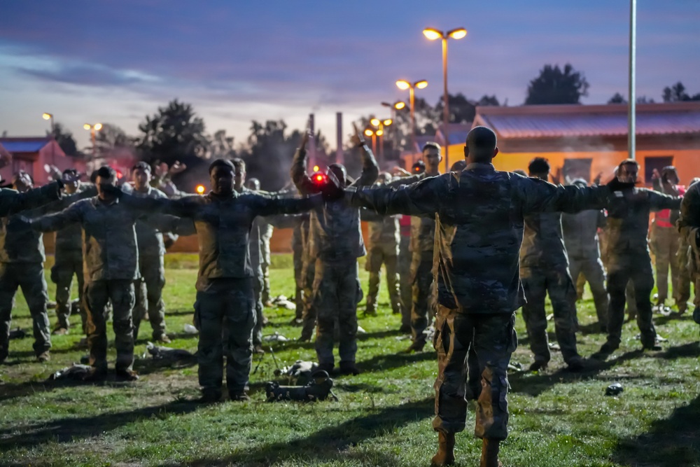 A military physical training or formation exercise is taking place outdoors at dusk or dawn. A leader facing away from the camera stands with arms outstretched, directing a large group of soldiers in camouflage uniforms who are spread out in formation on a grass field. Red flares or light sources are visible among the group, suggesting a structured drill or ceremony.