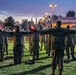 A military physical training or formation exercise is taking place outdoors at dusk or dawn. A leader facing away from the camera stands with arms outstretched, directing a large group of soldiers in camouflage uniforms who are spread out in formation on a grass field. Red flares or light sources are visible among the group, suggesting a structured drill or ceremony.