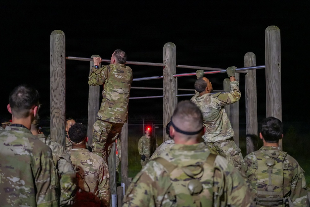 A group of military personnel in camouflage uniforms are conducting a nighttime physical training exercise at an outdoor obstacle course. Two soldiers are actively performing pull-ups on horizontal bars supported by wooden posts, while others observe and wait their turn. The scene is illuminated by artificial lighting against a completely dark background.