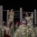 A group of military personnel in camouflage uniforms are conducting a nighttime physical training exercise at an outdoor obstacle course. Two soldiers are actively performing pull-ups on horizontal bars supported by wooden posts, while others observe and wait their turn. The scene is illuminated by artificial lighting against a completely dark background.