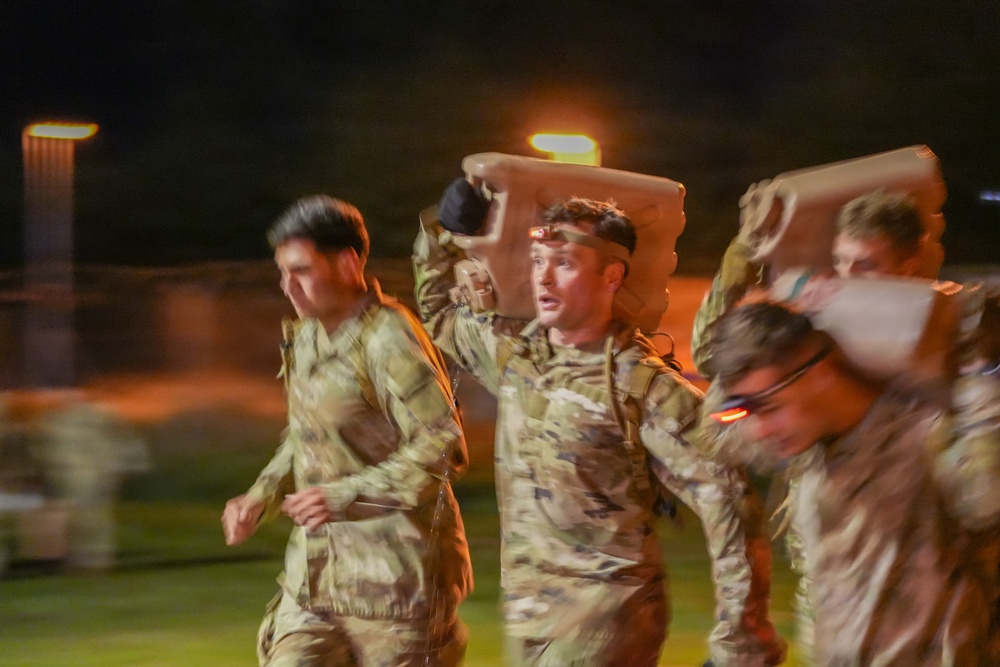 A group of military personnel in camouflage uniforms are running at night during what appears to be a training exercise or physical fitness event. At least one soldier is carrying a large object overhead, possibly a jerry can or equipment case, while wearing a headlamp. The image is motion-blurred, suggesting fast movement and dynamic action.