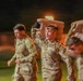 A group of military personnel in camouflage uniforms are running at night during what appears to be a training exercise or physical fitness event. At least one soldier is carrying a large object overhead, possibly a jerry can or equipment case, while wearing a headlamp. The image is motion-blurred, suggesting fast movement and dynamic action.