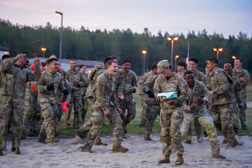 A large group of U.S. Army soldiers in MultiCam/OCP camouflage uniforms are celebrating enthusiastically in an outdoor area at dusk or dawn. The soldiers appear to be in high spirits, laughing, dancing, and cheering, suggesting the completion of a training exercise, competition, or significant milestone. The setting appears to be a military training area or base, likely in Europe based on the forested background and infrastructure.