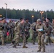 A large group of U.S. Army soldiers in MultiCam/OCP camouflage uniforms are celebrating enthusiastically in an outdoor area at dusk or dawn. The soldiers appear to be in high spirits, laughing, dancing, and cheering, suggesting the completion of a training exercise, competition, or significant milestone. The setting appears to be a military training area or base, likely in Europe based on the forested background and infrastructure.
