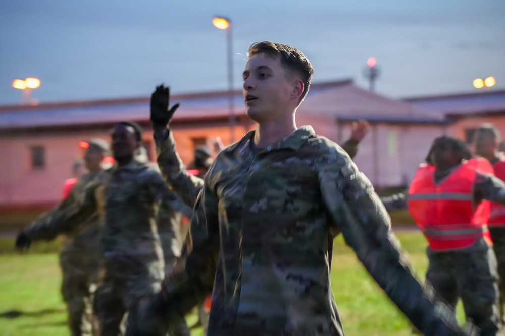 A group of military personnel in camouflage uniforms are performing physical training exercises outdoors during early morning or dusk hours. The foreground features a young male soldier with arms outstretched, appearing to lead or participate in a group exercise routine. Several other soldiers in the background, some wearing high-visibility orange safety vests, are performing similar movements.