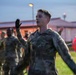 A group of military personnel in camouflage uniforms are performing physical training exercises outdoors during early morning or dusk hours. The foreground features a young male soldier with arms outstretched, appearing to lead or participate in a group exercise routine. Several other soldiers in the background, some wearing high-visibility orange safety vests, are performing similar movements.