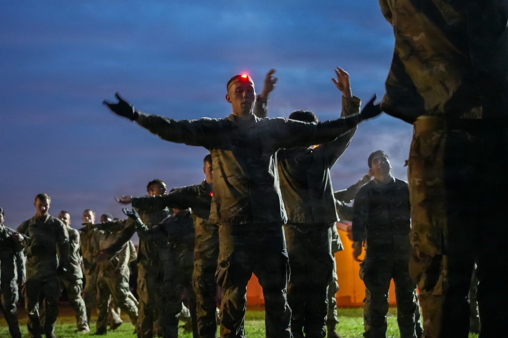 A group of military personnel in camouflage uniforms are conducting physical training exercises outdoors at dusk or dawn. The central figure wears a red LED headlamp and has arms outstretched, appearing to lead or demonstrate an exercise. Multiple rows of soldiers are visible performing synchronized movements in low-light conditions.
