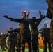 A group of military personnel in camouflage uniforms are conducting physical training exercises outdoors at dusk or dawn. The central figure wears a red LED headlamp and has arms outstretched, appearing to lead or demonstrate an exercise. Multiple rows of soldiers are visible performing synchronized movements in low-light conditions.