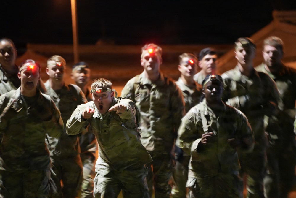 A group of military personnel in camouflage uniforms are conducting a nighttime physical training or formation run. Several individuals are wearing red LED headlamps, and one soldier in the foreground appears to be calling out cadence with an open mouth and pointing gesture. The scene is illuminated by ambient artificial lighting against a dark night sky.