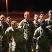 A group of military personnel in camouflage uniforms are conducting a nighttime physical training or formation run. Several individuals are wearing red LED headlamps, and one soldier in the foreground appears to be calling out cadence with an open mouth and pointing gesture. The scene is illuminated by ambient artificial lighting against a dark night sky.