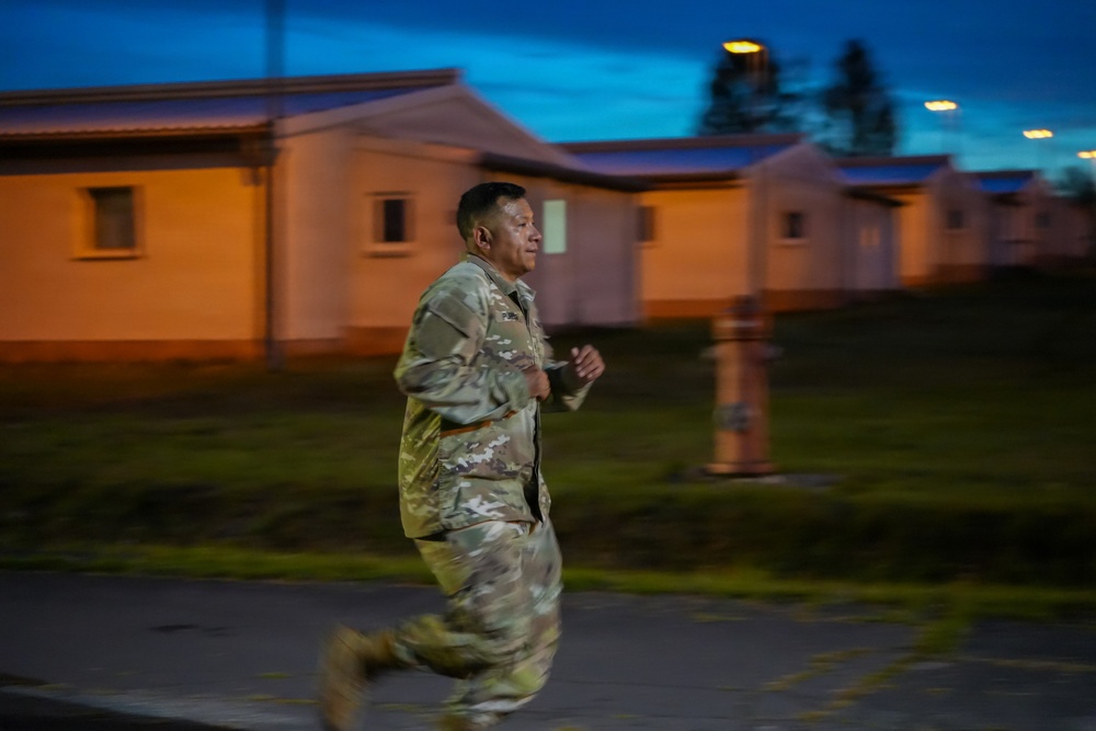 A soldier in camouflage uniform is running along a paved path on what appears to be a military installation during early morning or dusk hours. The background features low-profile barracks-style buildings illuminated by orange artificial lighting. Motion blur on the subject's lower body indicates significant movement speed.