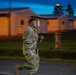 A soldier in camouflage uniform is running along a paved path on what appears to be a military installation during early morning or dusk hours. The background features low-profile barracks-style buildings illuminated by orange artificial lighting. Motion blur on the subject's lower body indicates significant movement speed.