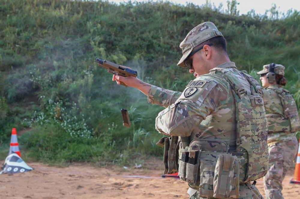 A U.S. Army Military Police (MP) soldier is actively firing a pistol at an outdoor shooting range, with a magazine visibly ejecting mid-air during a reload or malfunction drill. The soldier is wearing Operational Camouflage Pattern (OCP) uniform with full tactical gear including a plate carrier and magazine pouches. A second soldier in the background observes or awaits their turn, wearing hearing protection.