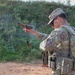 A U.S. Army Military Police (MP) soldier is actively firing a pistol at an outdoor shooting range, with a magazine visibly ejecting mid-air during a reload or malfunction drill. The soldier is wearing Operational Camouflage Pattern (OCP) uniform with full tactical gear including a plate carrier and magazine pouches. A second soldier in the background observes or awaits their turn, wearing hearing protection.