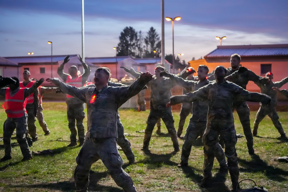 A group of military personnel in camouflage uniforms are performing physical training exercises outdoors during dusk or dawn. The soldiers are spread out on a grass field in a wide-stance position with arms extended, appearing to follow a leader at the front. The setting appears to be a military installation with buildings, street lights, and trees visible in the background.