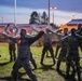 A group of military personnel in camouflage uniforms are performing physical training exercises outdoors during dusk or dawn. The soldiers are spread out on a grass field in a wide-stance position with arms extended, appearing to follow a leader at the front. The setting appears to be a military installation with buildings, street lights, and trees visible in the background.