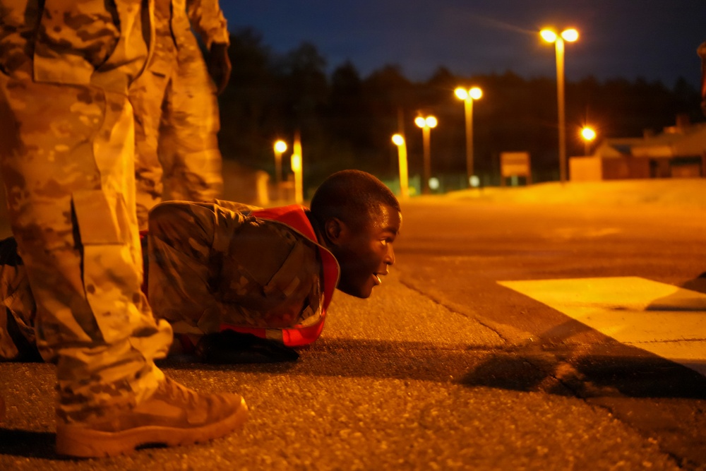 A soldier in camouflage uniform and an orange/red safety vest is performing push-ups on a paved surface at night. Another soldier in camouflage stands nearby, likely observing or supervising. The scene is illuminated by multiple street lights in what appears to be a military installation or training area.
