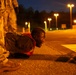 A soldier in camouflage uniform and an orange/red safety vest is performing push-ups on a paved surface at night. Another soldier in camouflage stands nearby, likely observing or supervising. The scene is illuminated by multiple street lights in what appears to be a military installation or training area.