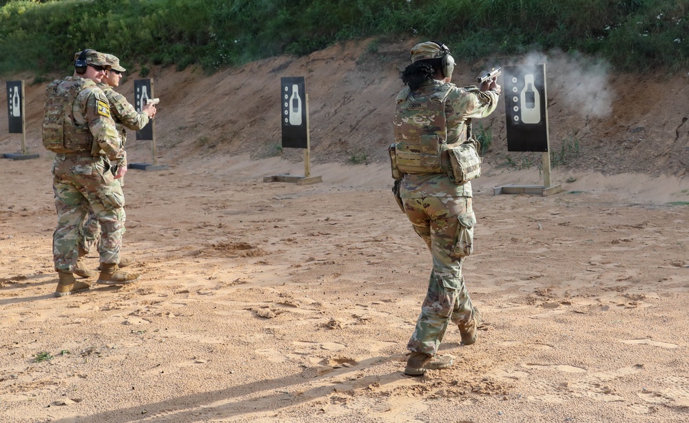 Two U.S. Army soldiers in OCP (Operational Camouflage Pattern) uniforms are conducting pistol qualification or training at an outdoor firing range. One soldier is actively firing at a silhouette target, with visible muzzle smoke, while a second soldier observes or awaits their turn. The range features multiple black silhouette targets mounted on wooden posts against a sandy berm backdrop.