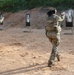 Two U.S. Army soldiers in OCP (Operational Camouflage Pattern) uniforms are conducting pistol qualification or training at an outdoor firing range. One soldier is actively firing at a silhouette target, with visible muzzle smoke, while a second soldier observes or awaits their turn. The range features multiple black silhouette targets mounted on wooden posts against a sandy berm backdrop.