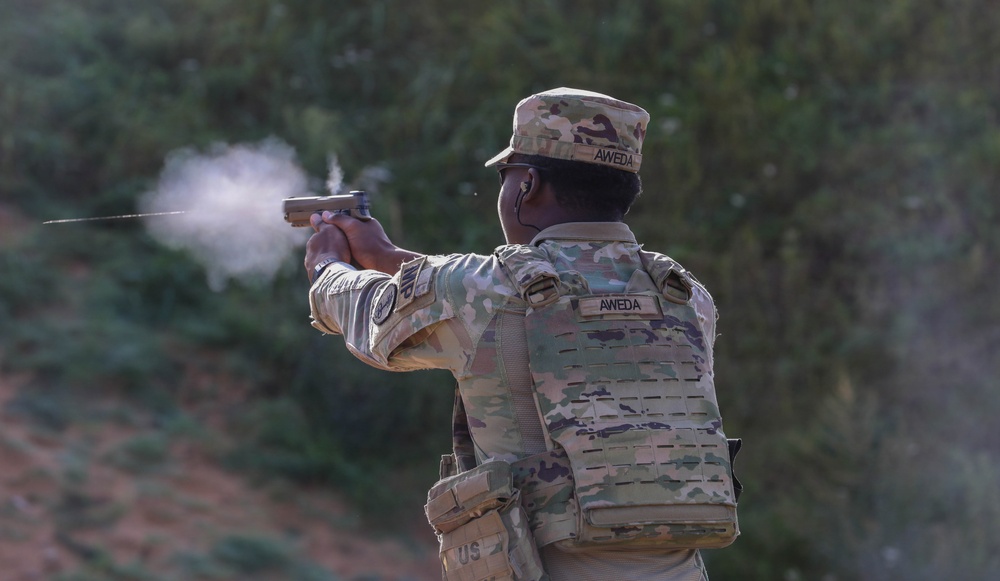 A U.S. Army soldier identified by the name tape 'AWEDA' is firing a pistol at a shooting range. The soldier is wearing Operational Camouflage Pattern (OCP) uniform with a tactical plate carrier vest and patrol cap. Visible gun smoke/muzzle blast indicates the weapon has just been discharged.
