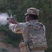 A U.S. Army soldier identified by the name tape 'AWEDA' is firing a pistol at a shooting range. The soldier is wearing Operational Camouflage Pattern (OCP) uniform with a tactical plate carrier vest and patrol cap. Visible gun smoke/muzzle blast indicates the weapon has just been discharged.