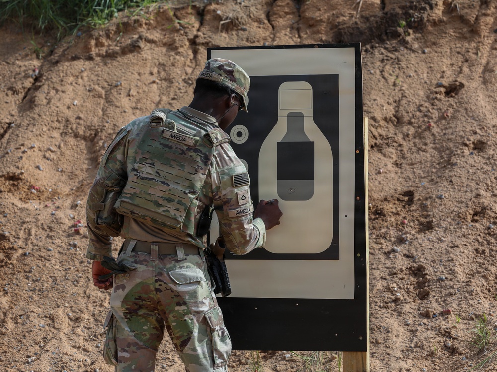 A U.S. Army soldier in OCP (Operational Camouflage Pattern) uniform is standing at a shooting range target board, appearing to inspect or mark the target after a firing exercise. The soldier, identified by name tape as 'AWEDA,' is wearing a plate carrier vest and has a pistol holstered at the hip. The setting is an outdoor range with a sandy berm backdrop.