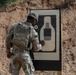 A U.S. Army soldier in OCP (Operational Camouflage Pattern) uniform is standing at a shooting range target board, appearing to inspect or mark the target after a firing exercise. The soldier, identified by name tape as 'AWEDA,' is wearing a plate carrier vest and has a pistol holstered at the hip. The setting is an outdoor range with a sandy berm backdrop.