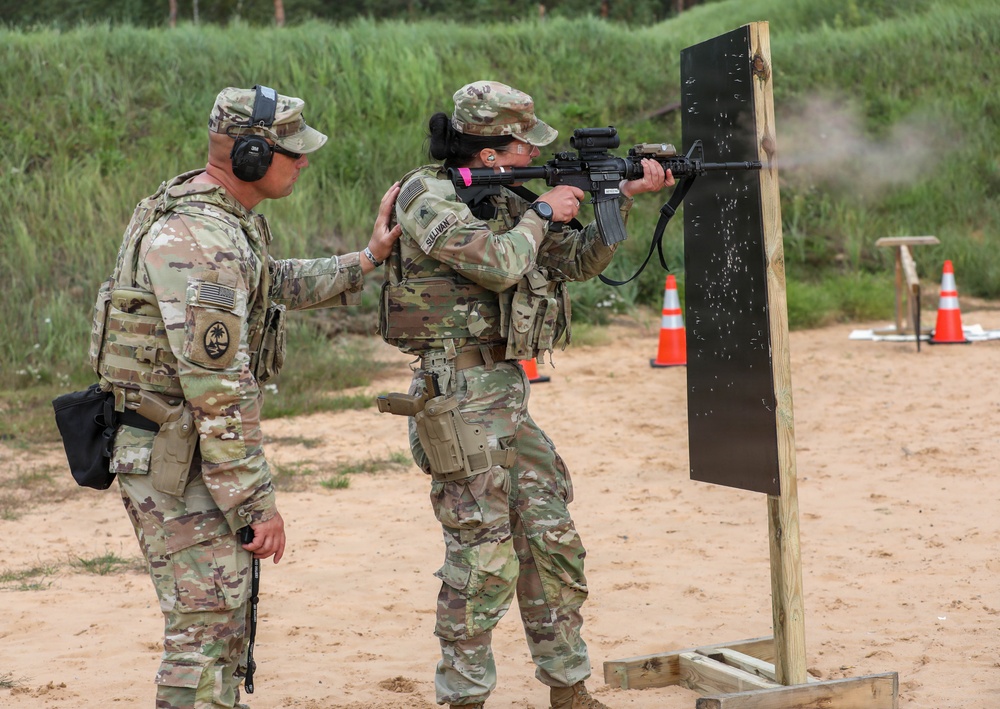Two military personnel in OCP (Operational Camouflage Pattern) uniforms are at an outdoor shooting range. One soldier is actively firing an M4/M16-style rifle at a black target board mounted on a wooden stand, while a second soldier provides hands-on instruction or guidance. The setting appears to be a sand-based military training range with safety cones and a vegetated berm in the background.