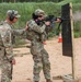 Two military personnel in OCP (Operational Camouflage Pattern) uniforms are at an outdoor shooting range. One soldier is actively firing an M4/M16-style rifle at a black target board mounted on a wooden stand, while a second soldier provides hands-on instruction or guidance. The setting appears to be a sand-based military training range with safety cones and a vegetated berm in the background.