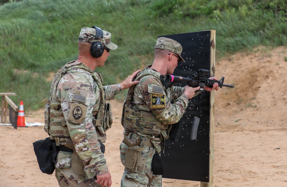 Two military personnel in camouflage uniforms are at an outdoor shooting range. One soldier is actively firing a rifle at a black target board while another observes and provides guidance with a hand on the shooter's shoulder. The target board shows numerous bullet impact marks, indicating active training.