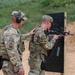 Two military personnel in camouflage uniforms are at an outdoor shooting range. One soldier is actively firing a rifle at a black target board while another observes and provides guidance with a hand on the shooter's shoulder. The target board shows numerous bullet impact marks, indicating active training.
