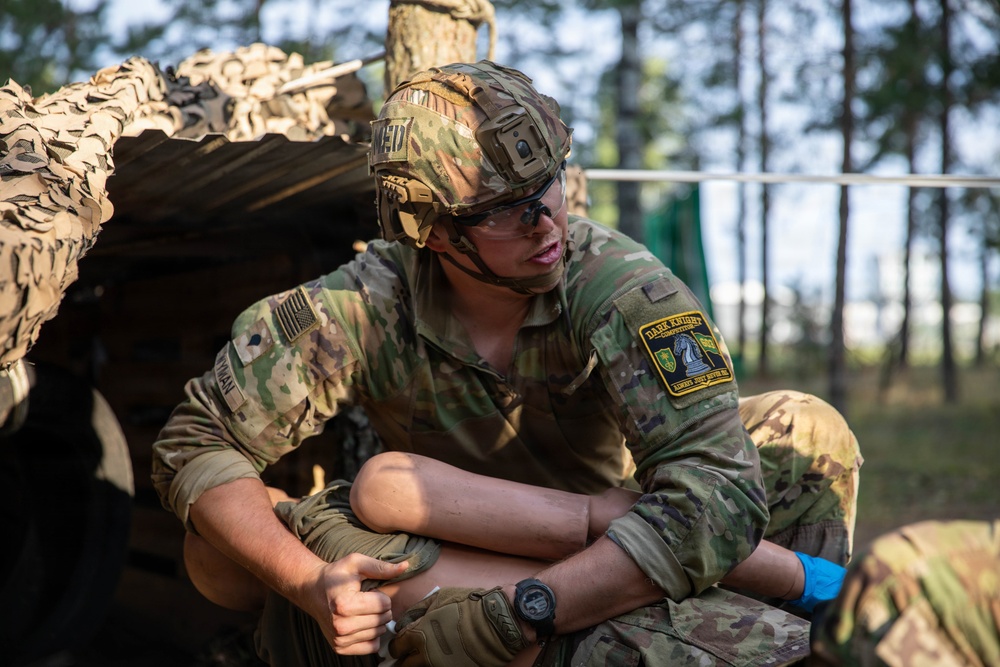 A U.S. Army soldier in full multicam combat uniform is crouched or seated near a camouflage-netted position in a wooded area. The soldier is wearing a tactical helmet with a mounted device, protective eyewear, and blue medical gloves, suggesting participation in a medical or combat training exercise. A 'Dark Knight Competition' patch is visible on the right shoulder, indicating this is likely a military skills competition event.