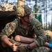 A U.S. Army soldier in full multicam combat uniform is crouched or seated near a camouflage-netted position in a wooded area. The soldier is wearing a tactical helmet with a mounted device, protective eyewear, and blue medical gloves, suggesting participation in a medical or combat training exercise. A 'Dark Knight Competition' patch is visible on the right shoulder, indicating this is likely a military skills competition event.