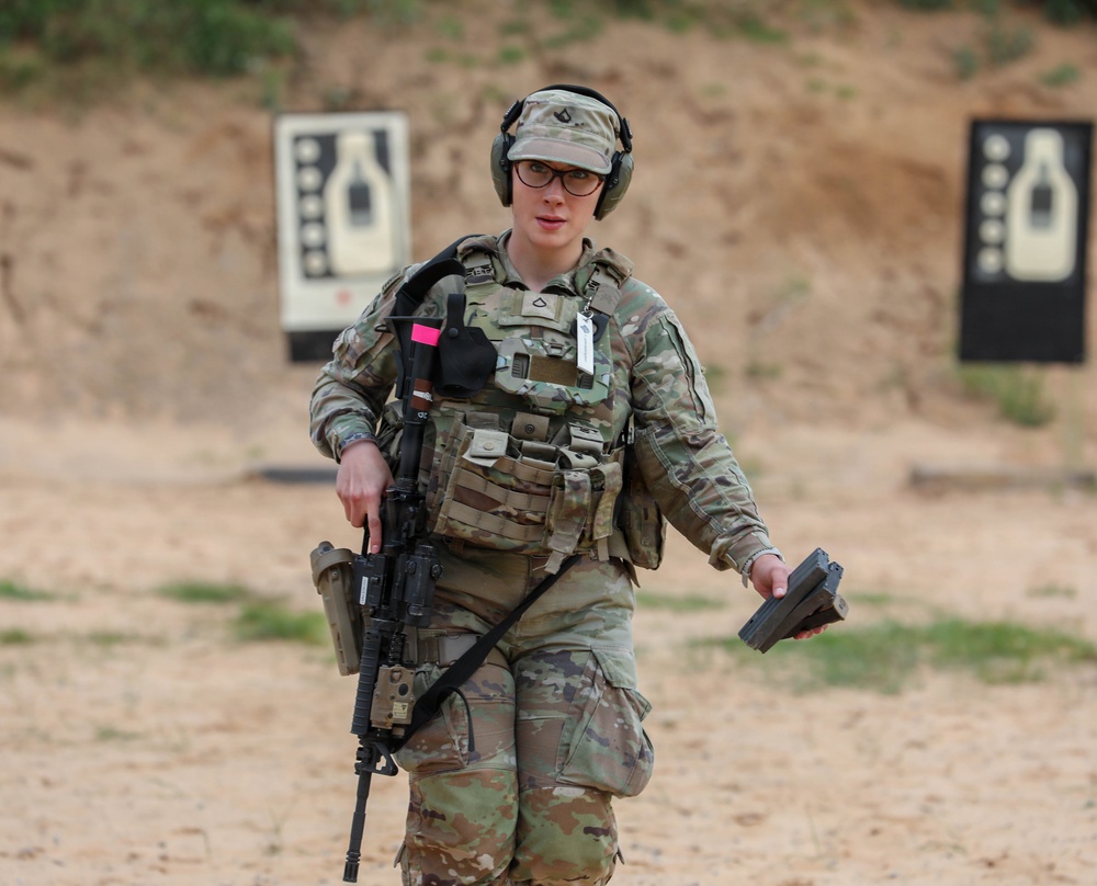 A soldier in U.S. Army OCP (Operational Camouflage Pattern) uniform is walking on a firing range, carrying an M4-style carbine slung across the body and holding what appears to be a pistol magazine in the right hand. The soldier is equipped with a plate carrier vest, hearing protection headset, ballistic glasses, and a patrol cap. Silhouette target boards are visible in the background, indicating an active weapons qualification or training exercise.