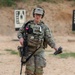 A soldier in U.S. Army OCP (Operational Camouflage Pattern) uniform is walking on a firing range, carrying an M4-style carbine slung across the body and holding what appears to be a pistol magazine in the right hand. The soldier is equipped with a plate carrier vest, hearing protection headset, ballistic glasses, and a patrol cap. Silhouette target boards are visible in the background, indicating an active weapons qualification or training exercise.