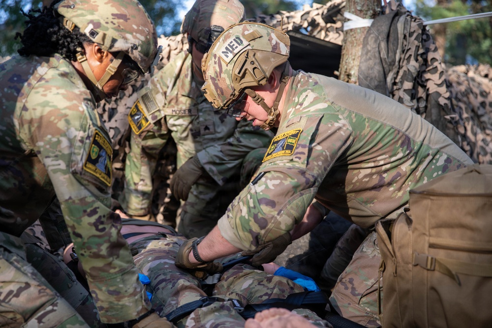 Military medical personnel in OCP (Operational Camouflage Pattern) uniforms are performing tactical combat casualty care (TCCC) on a simulated or actual casualty lying on a litter/stretcher. At least three soldiers are actively involved, with one wearing a helmet marked 'MED' indicating a medic role. The scene appears to take place under camouflage netting in a field environment.