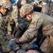 Military medical personnel in OCP (Operational Camouflage Pattern) uniforms are performing tactical combat casualty care (TCCC) on a simulated or actual casualty lying on a litter/stretcher. At least three soldiers are actively involved, with one wearing a helmet marked 'MED' indicating a medic role. The scene appears to take place under camouflage netting in a field environment.