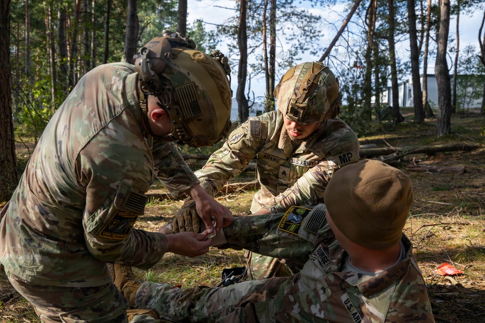Three U.S. Army soldiers in full combat gear are conducting tactical combat casualty care (TCCC) training in a forested environment. Two soldiers wearing helmets with night vision mounts are administering first aid to a third soldier seated on the ground. The setting appears to be a field training exercise based on the woodland surroundings and a structure visible in the background.