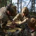 Three U.S. Army soldiers in full combat gear are conducting tactical combat casualty care (TCCC) training in a forested environment. Two soldiers wearing helmets with night vision mounts are administering first aid to a third soldier seated on the ground. The setting appears to be a field training exercise based on the woodland surroundings and a structure visible in the background.