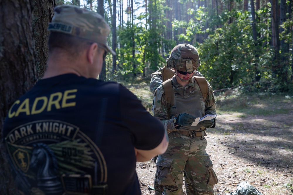 Two individuals are in a forested outdoor setting, one wearing a black 'CADRE - Dark Knight Competition' t-shirt and a camouflage cap, and the other in full military OCP (Operational Camouflage Pattern) uniform with helmet, body armor, and a rucksack. The uniformed soldier appears to be writing or reviewing notes on a small notepad. This appears to be a military competition or training event, likely a land navigation or skills assessment exercise.