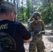 Two individuals are in a forested outdoor setting, one wearing a black 'CADRE - Dark Knight Competition' t-shirt and a camouflage cap, and the other in full military OCP (Operational Camouflage Pattern) uniform with helmet, body armor, and a rucksack. The uniformed soldier appears to be writing or reviewing notes on a small notepad. This appears to be a military competition or training event, likely a land navigation or skills assessment exercise.